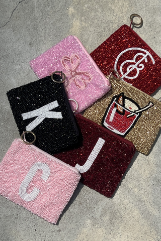 A small black beaded pouch with an embroidered green olive and toothpick lies on wrinkled, shiny white satin fabric. The pouch has a zipper and a silver keyring attachment.