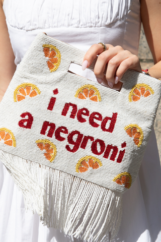 A person in a white dress holds a beaded white clutch with orange slice designs and red text that reads i need a negroni. The bottom of the clutch has white fringe.