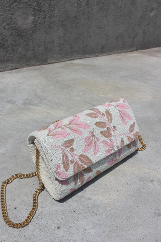 A small rectangular purse with a gold chain strap lies on cream satin fabric. The purse features a white, gold, and olive green beaded geometric and floral pattern.