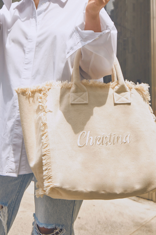 A person in a white shirt and ripped jeans holds a beige fringed tote bag embroidered with the name Christina in white script, standing outdoors on a sunny day.