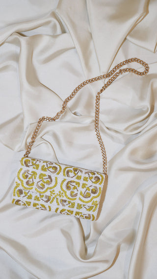 A small rectangular purse with a gold chain strap lies on cream satin fabric. The purse features a white, gold, and olive green beaded geometric and floral pattern.
