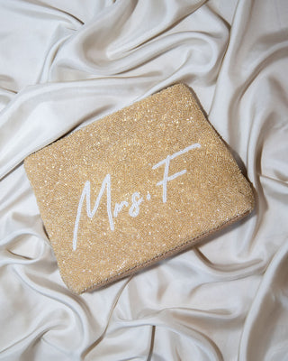 A gold glittery clutch purse with “Mrs. F” in white script lies on smooth, cream-colored satin fabric, creating a soft and elegant background.