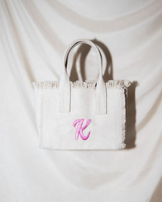 A white canvas tote bag with frayed edges and two handles hangs against a white fabric backdrop. The bag features a pink embroidered letter K on the front.