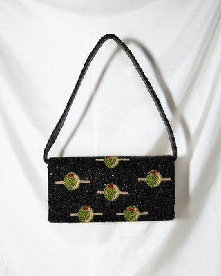 A black rectangular beaded handbag with a single strap, decorated with embroidered green olives on toothpicks, is displayed against a draped white fabric background.
