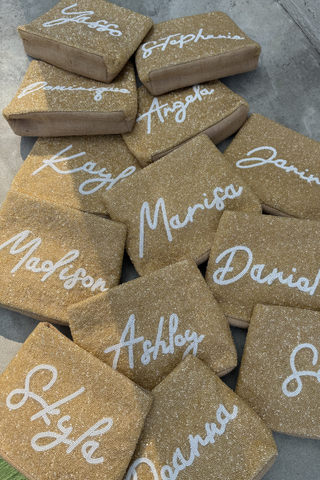 A gold glittery clutch purse with “Mrs. F” in white script lies on smooth, cream-colored satin fabric, creating a soft and elegant background.